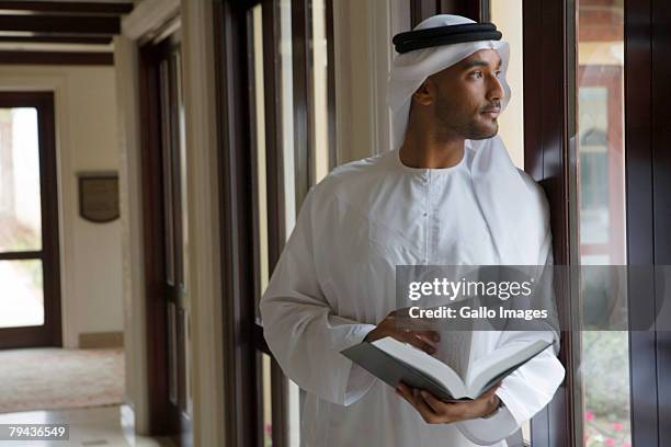 arab man looking out window, holding book - arabic book stock pictures, royalty-free photos & images