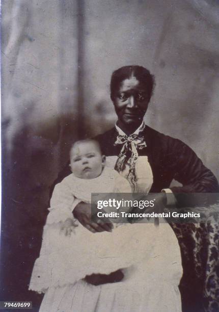 Portrait of African American woman wearing an apron, sitting and holding a white infant in a white dress on her lap, circa 1875. USA