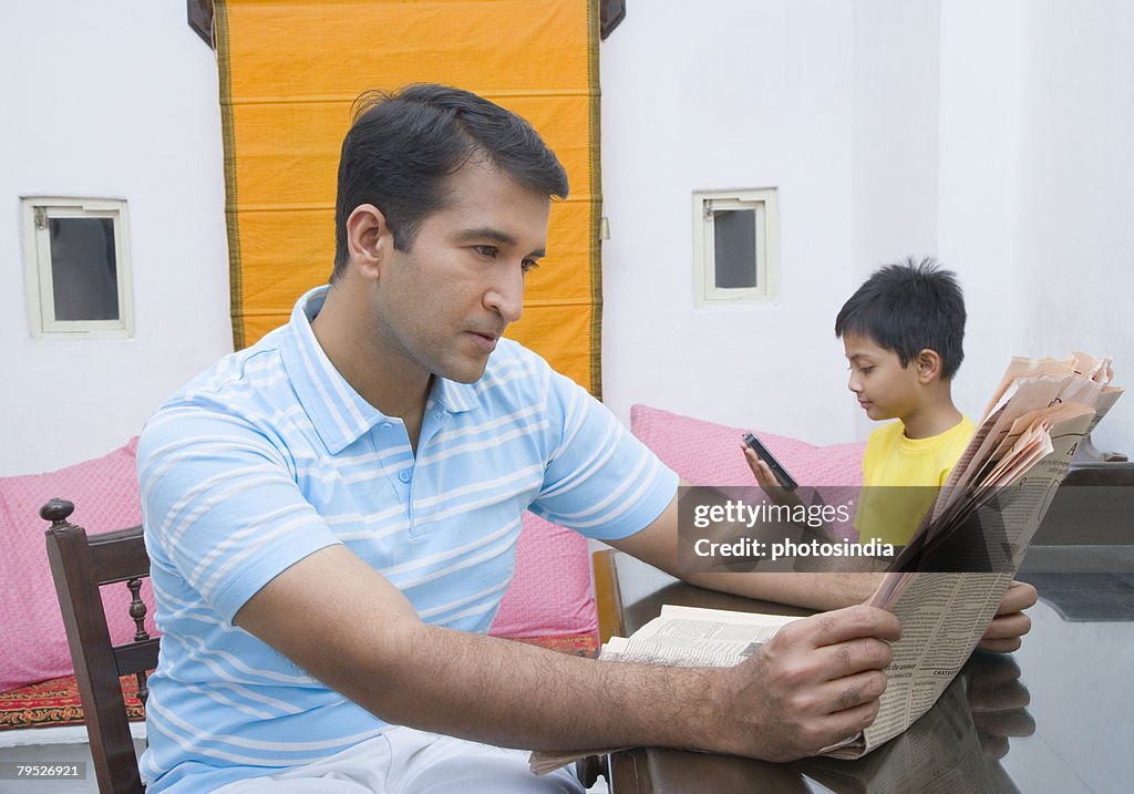 Mid adult man reading a newspaper with his son holding a mobile phone in the background