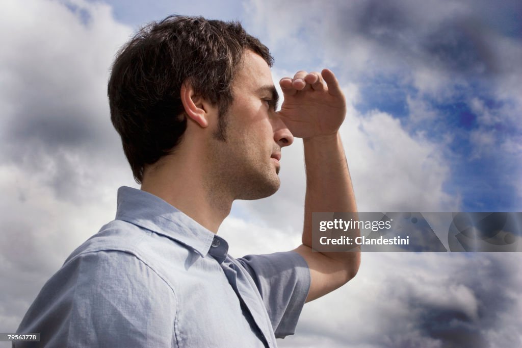 Young man against cloudy sky, shielding eyes, side view, close-up