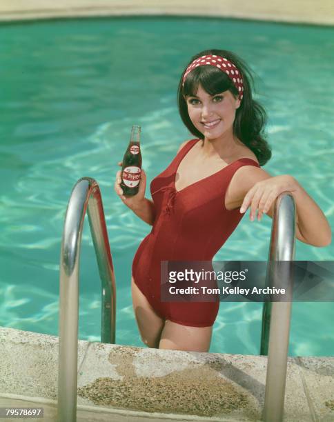 An unidentified model in a red swimsuit and headband smiles as she poses on a swimming pool ladder with bottle of Dr. Pepper soda in one hand, June...