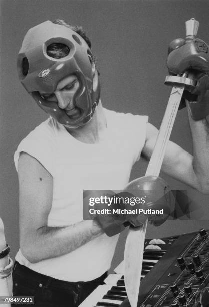 English musician, keyboard player and composer Dave Stewart pictured wearing boxing gloves and helmet as he attempts to insert a sword in a keyboard...