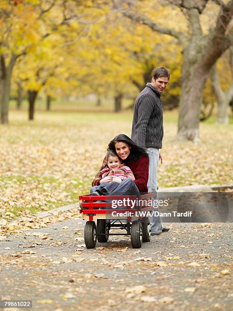 father pulling mother and daughter in wagon - bollerwagen mann stock-fotos und bilder