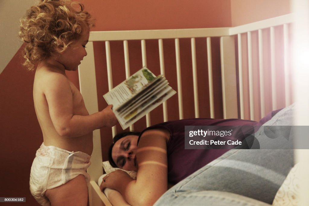 Toddler reading mother a book while she is asleep in a cot
