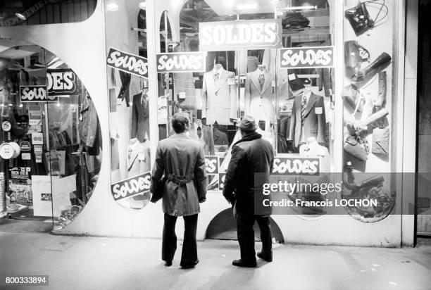 Deux hommes regardent la vitrine d'un magasin d'habillement en période de soldes en février 1976 à Paris, France.