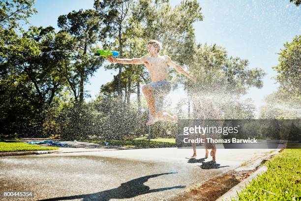 teen boy with squirt gun jumping through spray from fire hydrant on sunny summer afternoon - teen boy shorts stockfoto's en -beelden