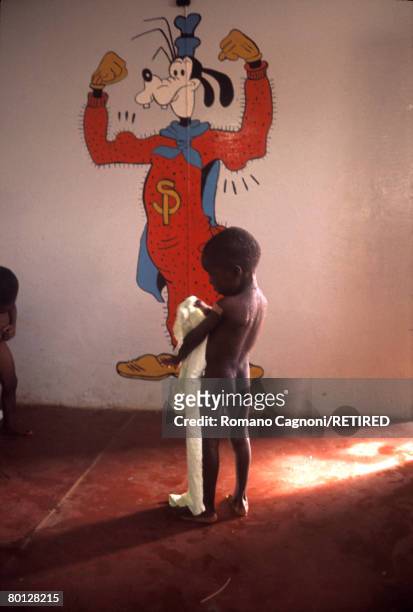 An emaciated child stands in front of a drawing of Goofy in Biafra, a secessionist state in southern Nigeria, circa 1970.