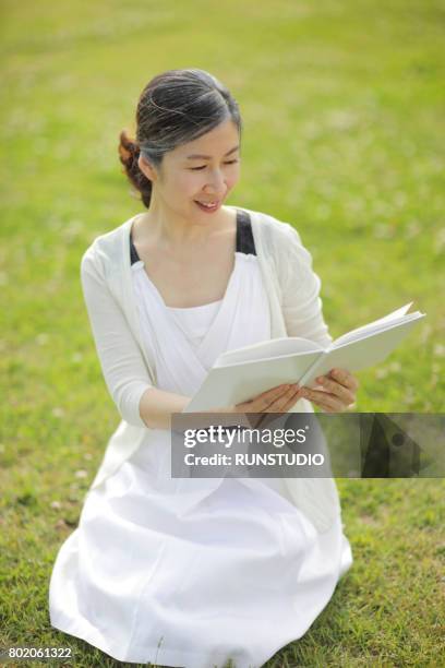 senior woman sitting on grass, reading book - fersensitz stock-fotos und bilder