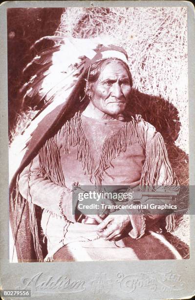 Portrait of Chiricahua Apache Geronimo as he sits next to a haybale, a revolver in one hand on his lap, early 1900s.