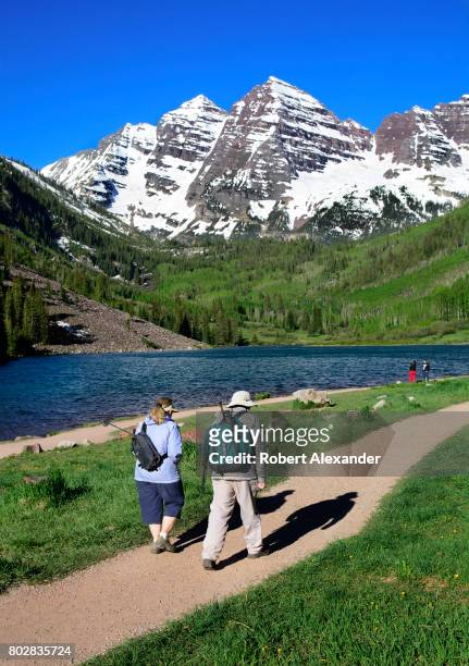 Two hikers walk past Maroon Lake with the Maroon Bells in the background. The Maroon Bells, near Aspen, Colorado, are two peaks in the Elk Mountains...