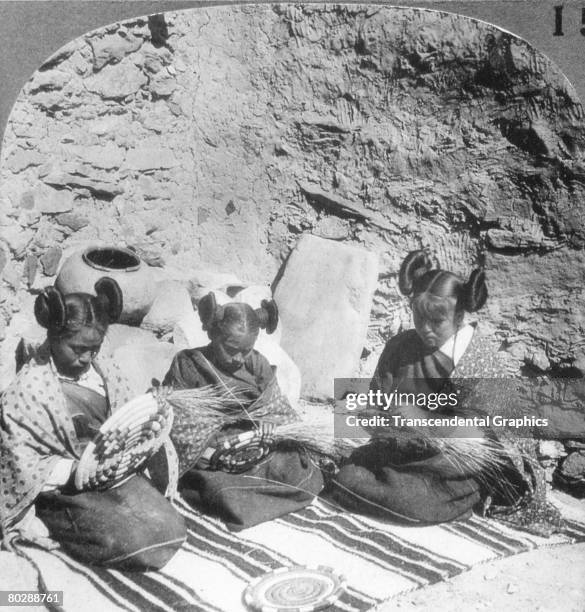 Three young Hopi women weave baskets on a blanket near a rough-hewn stone wall, early twentieth century. The 'squash-blossom' or 'butterfly whorl'...