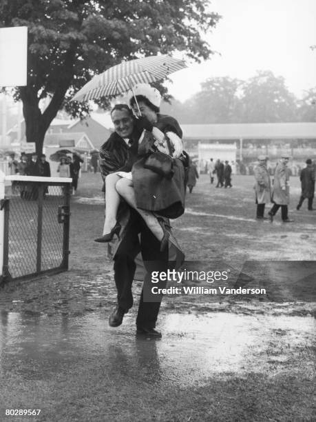 Man carries a woman over a muddy puddle as rain washes out the third day of racing at the Royal Ascot race meeting, 18th June 1964.