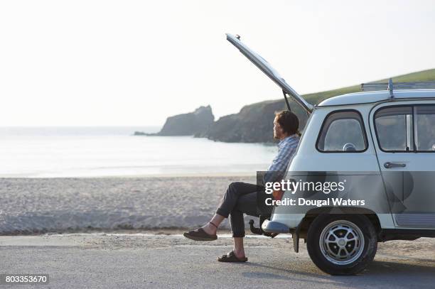 man sitting in boot of retro car at beach. - oldtimerauto stockfoto's en -beelden