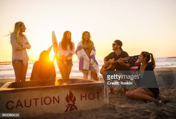 multi ethnic group of young people, spending time watching sunset at the beach in san diego, playing guitar and warming themselves by the fire - costa diego imagens e fotografias de stock