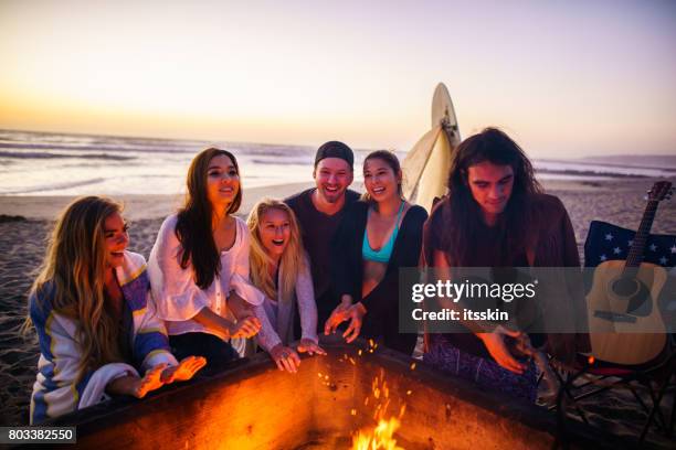 multi ethnic group of young people, spending time watching sunset at the beach in san diego, playing guitar and warming themselves by the fire - bonfire beach stock pictures, royalty-free photos & images