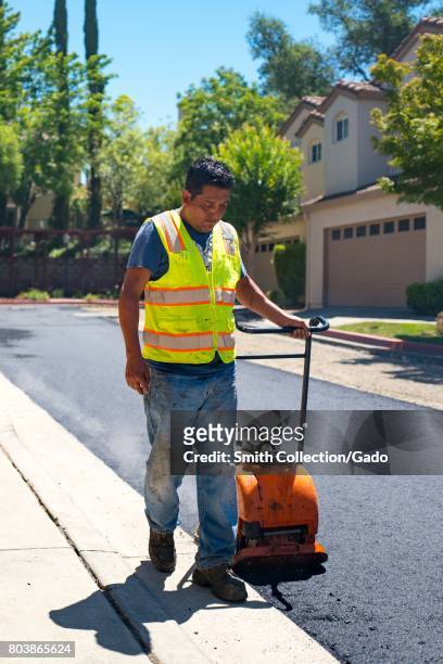 An employee of American Asphalt pulls a manual compactor across a newly laid road surface during a road construction and resurfacing project in the...