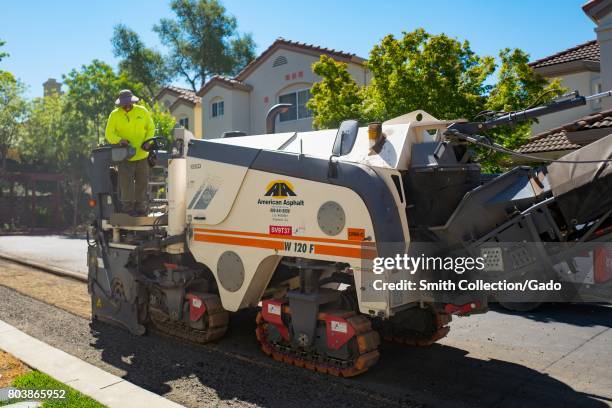 Construction worker for American Asphalt, wearing a bright yellow high visibility vest, steers a machine as it removes asphalt during a road...