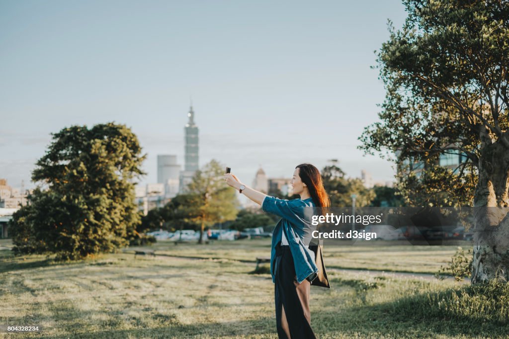 Joyful young lady taking pictures of beautiful scenics in park with smartphone during sunset