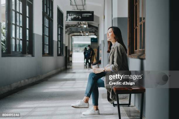 female asian student enjoying coffee and resting on bench in college - college campus stockfoto's en -beelden