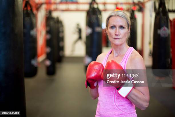 mature female boxer stands next to punch bag in gym with boxing gloves on - roze handschoen stockfoto's en -beelden