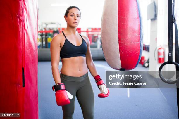 female boxer stands next to punch bag in gym with boxing gloves on - gants de sport photos et images de collection