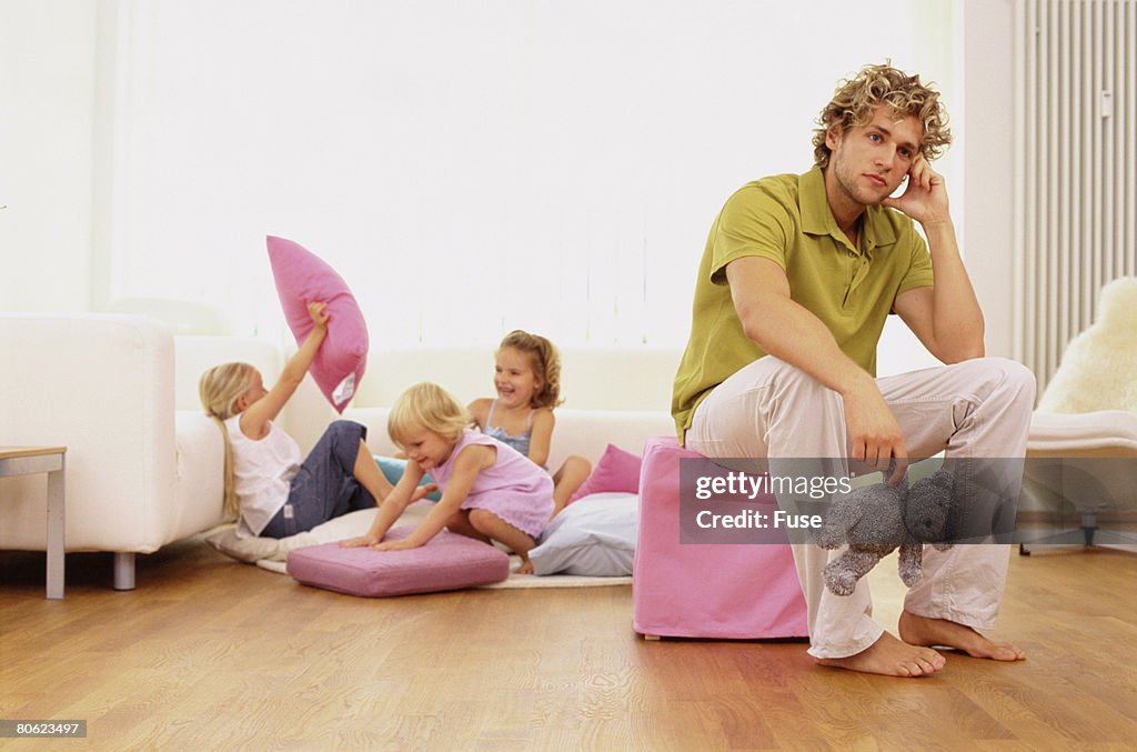 Father sitting on stool, three sisters having pillow fight