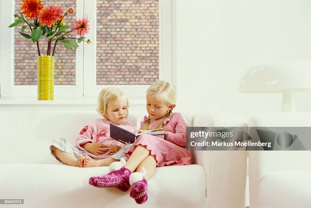 Two sisters sitting on sofa, reading a childrens book