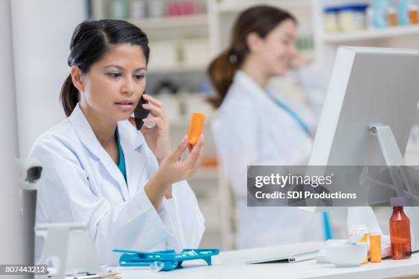young pharmacist studies prescription bottle during phone conversation - prescription drugs dangers stock pictures, royalty-free photos & images