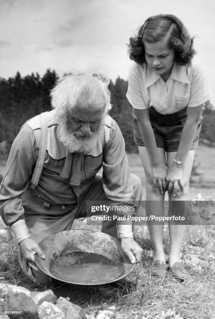 People Social History. pic: circa 1940's. Black Hills, South Dakota, USA. An old gold prospector seen prospecting for gold with an interested spectator alongside.