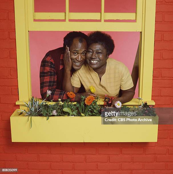 Promotional portrait of American actors John Amos and Esther Rolle as they pose behind a flowerbox on a windowframe for the television show 'Good...