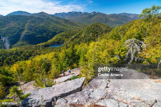 Young couple clambering up rocky slope overlooking New England's Adirondack Park in autumn, upstate New York, USA.