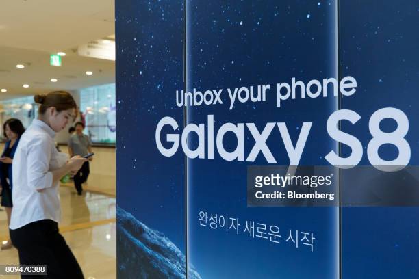 Woman using a smartphone walks past an advertisement for the Samsung Electronics Co. Galaxy S8 smartphone at the company's D'light flagship store in...