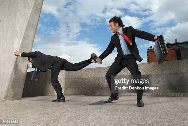 businessman pulling colleagues leg on windy roof - pull foto e immagini stock