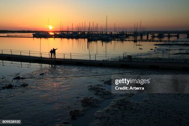 Couple on jetty silhouetted against sunset and sailing yachts in marina at El Rompido, coastal borough of Cartaya, province of Huelva, AndalucÕa,...