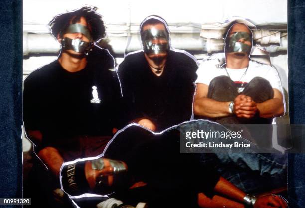 Vocalist Zack de la Rocha, bassist Tim Comerford, and drummer Brad Wilk are seated on the floor behind recumbent guitarist Tom Morello, all bound,...