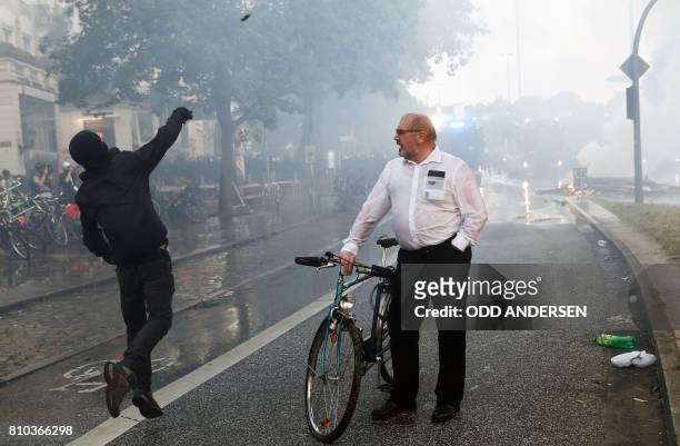 Man on his bike looks at a masked protester throwning a stone at riot police using water cannon on July 7, 2017 in Hamburg, northern Germany, where...