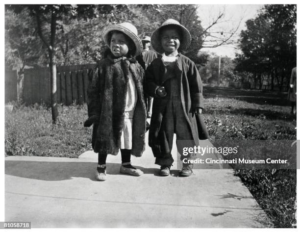 Portrait of a pair of children, both in straw hats and warm jackets , stand on a paved pathway in an unidentified public park, Kentucky, early 20th...