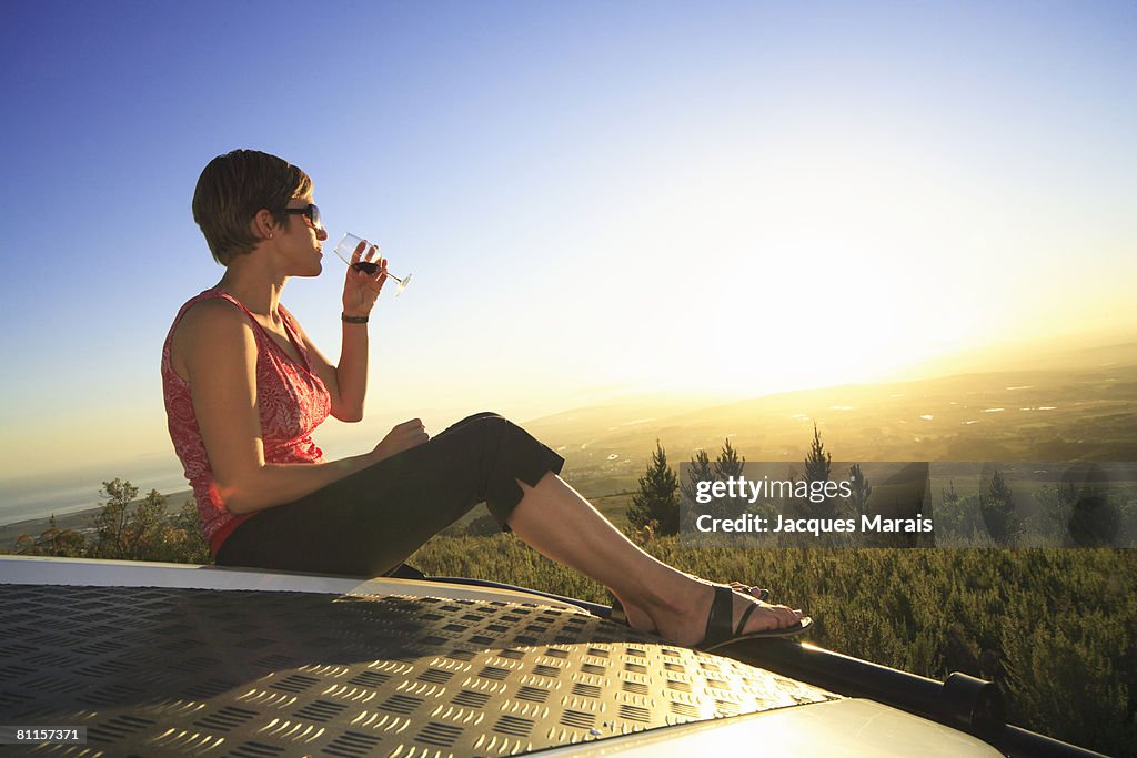 Woman sitting on a 4x4 vehicle tasting wine as sun sets in the Cape Winelands above Stellenbosch, Western Cape Province, South Africa