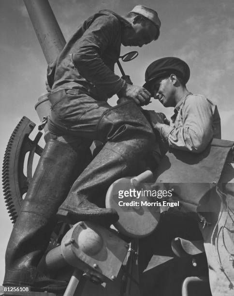 Unidentified American officer looks through the sight of a deck-mounted gun, assisted by a sailor in thigh-high rubber boots, 1940s.