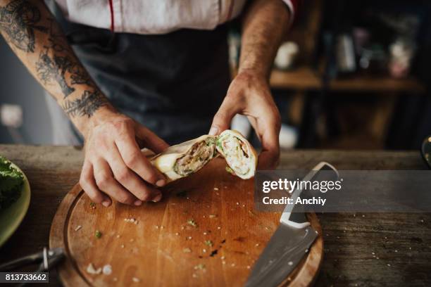 close up of tattooed chef's hands holding a wrap sandwich - food on cutting board stock pictures, royalty-free photos & images