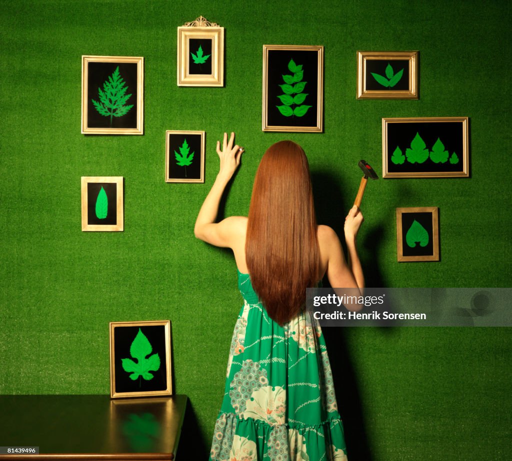 Green room with a woman hanging pictures of leaves on the wall.