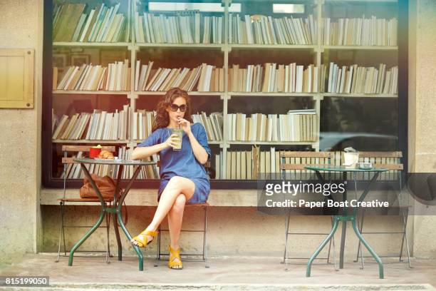 attractive young woman sipping juice sitting at an outdoor cafe table in front of a booktore with books on shelves behind her - sidewalk cafe stock pictures, royalty-free photos & images