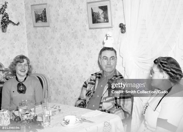 Family sits at a dinner table in a suburban home, the father goofing around by placing a toy snowman on his head, the daughter laughing at her...