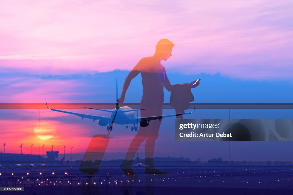 Silhouette of a guy checking his smartphone ready to departure on a airport at sunset with double exposure effect during a travel with trolley suitcase using application technology to get information about the trip.