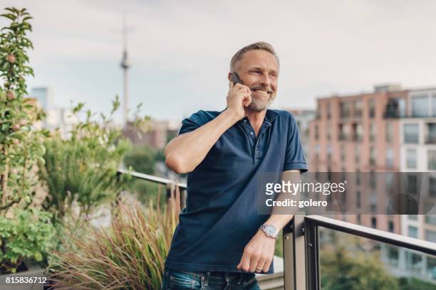 ベルリンのバルコニーで携帯電話で話しているハンサムなひげを生やした midaged 男 - ポロシャツ ストックフォトと画像
