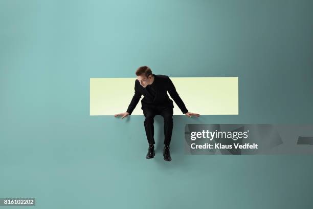 man sitting on the edge of rectangular opening in coloured wall, looking down - changing appearance photos et images de collection