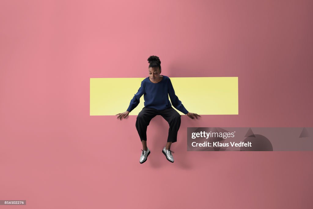 Woman sitting on the edge of rectangular opening in coloured wall and looking down