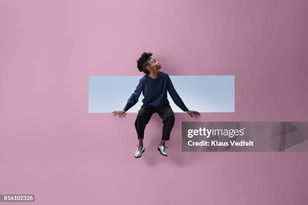 woman sitting on the edge of rectangular opening in coloured wall and looking up - purple pants stock-fotos und bilder