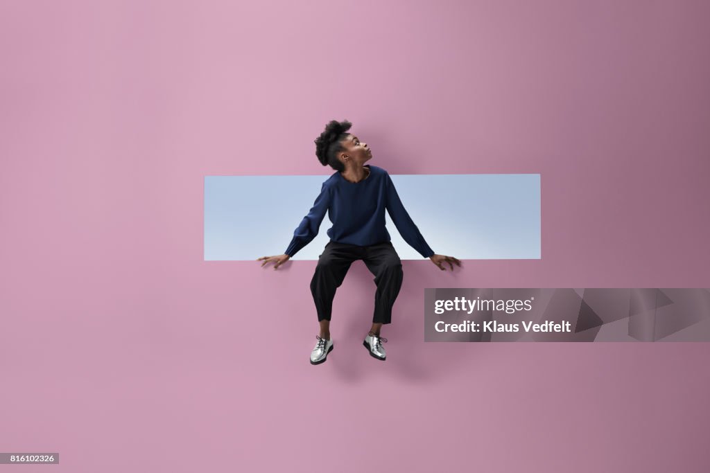 Woman sitting on the edge of rectangular opening in coloured wall and looking up