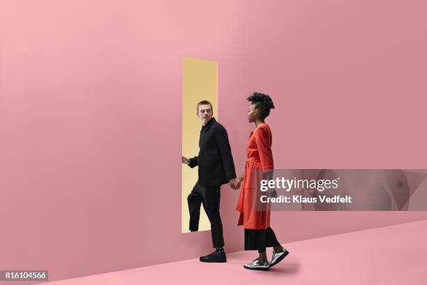 woman & man holding hands, approaching rectangular opening in coloured wall - gele overjas stockfoto's en -beelden